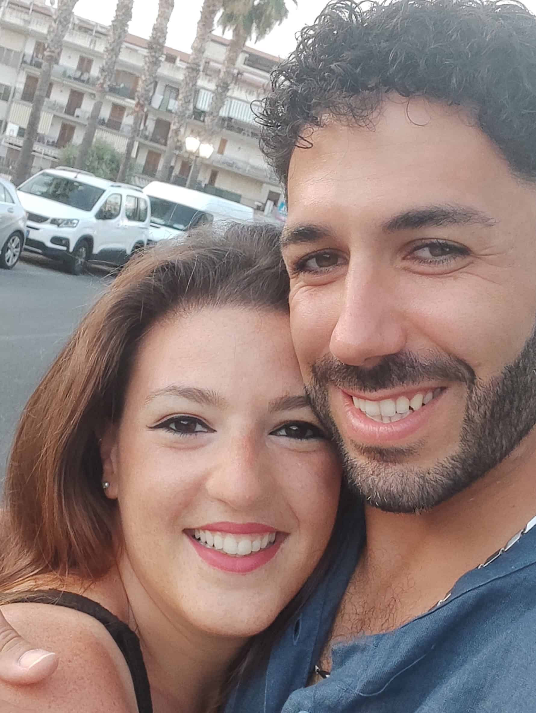 Due persone sorridenti in un selfie, ambiente urbano sullo sfondo.